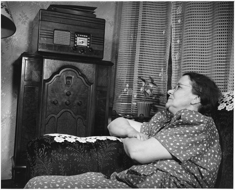 A woman in a 1940s parlor reclining beside a console radio, listening intently.